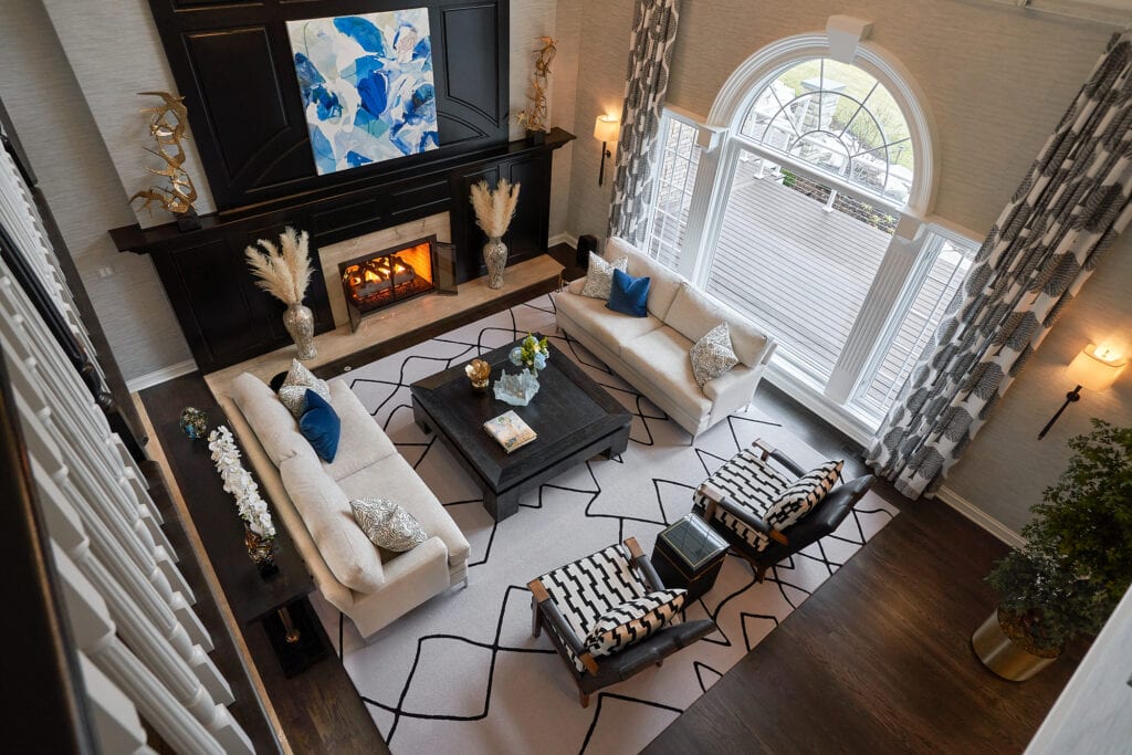 Living room with fireplace, viewed from above, patterned chairs, and large windows.
